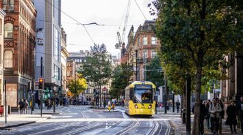 A yellow tram travels through a busy street in Manchester, surrounded by pedestrians and historic buildings