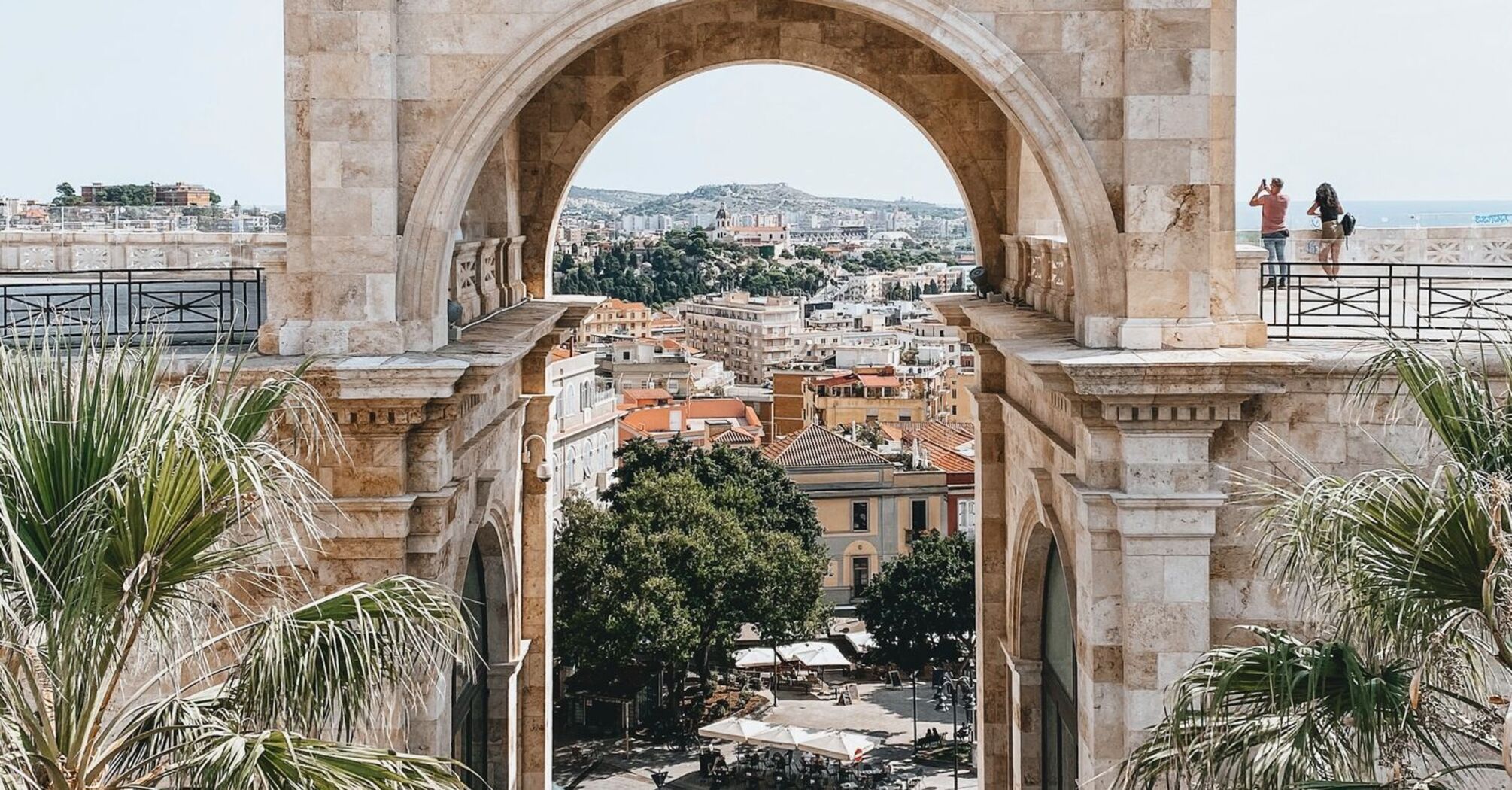 A historic stone archway in Cagliari, Sardinia, framing a scenic city view with palm trees and a lively square below