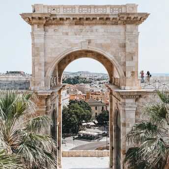 A historic stone archway in Cagliari, Sardinia, framing a scenic city view with palm trees and a lively square below