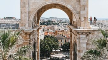 A historic stone archway in Cagliari, Sardinia, framing a scenic city view with palm trees and a lively square below