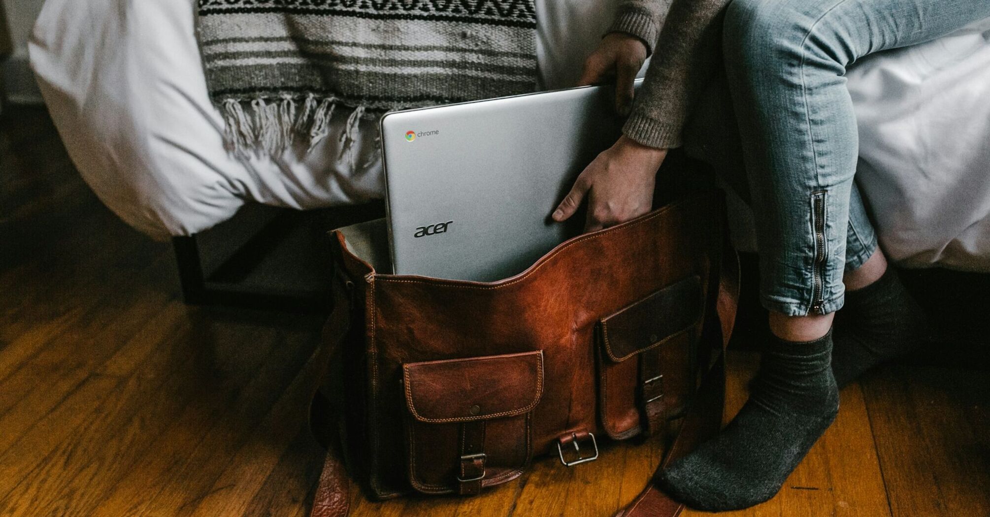 Person placing an Acer Chromebook into a leather travel bag beside a bed