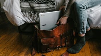Person placing an Acer Chromebook into a leather travel bag beside a bed