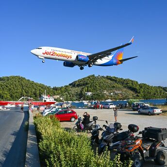 Jet2holidays airplane flying low over a coastal road near a populated area with parked vehicles and scenic green hills