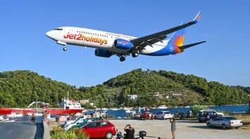 Jet2holidays airplane flying low over a coastal road near a populated area with parked vehicles and scenic green hills
