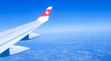A Swiss International Air Lines aircraft wing with the airline's red and white logo, flying high above a vast landscape under a clear blue sky