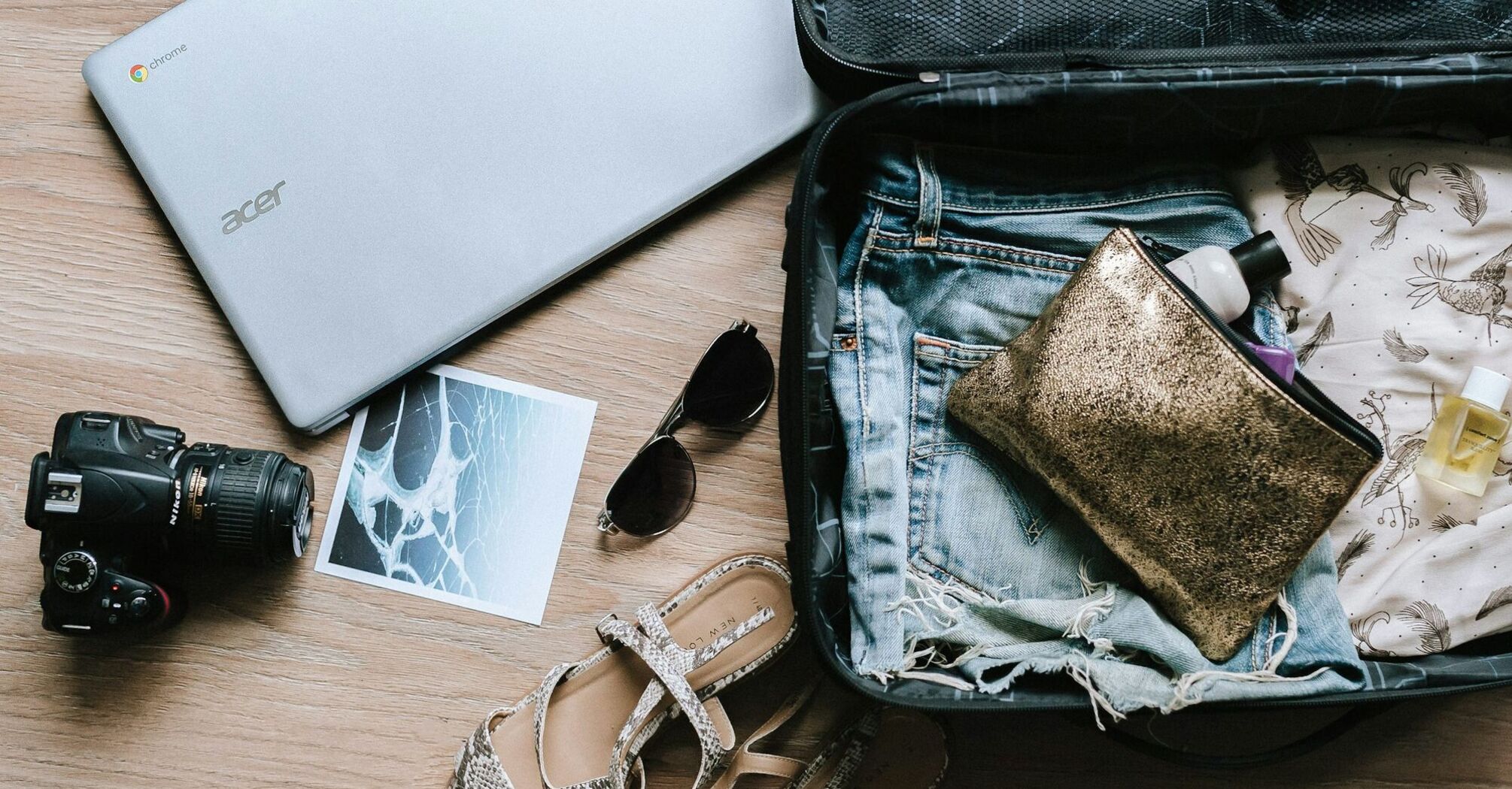 A packed suitcase with travel essentials including a laptop, camera, sunglasses, toiletries, and summer clothing on a wooden surface