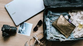 A packed suitcase with travel essentials including a laptop, camera, sunglasses, toiletries, and summer clothing on a wooden surface