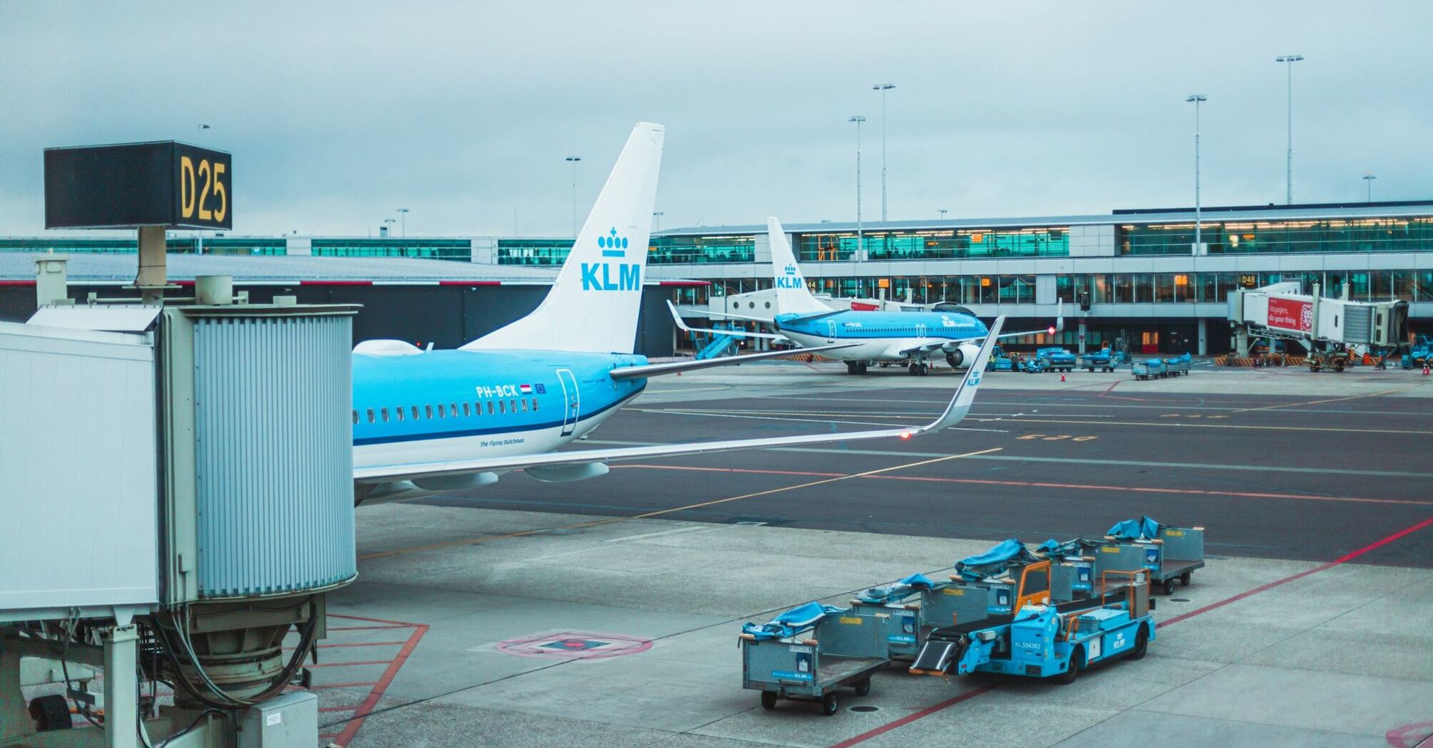 Two KLM airplanes parked at the airport gate with baggage handling equipment on the tarmac