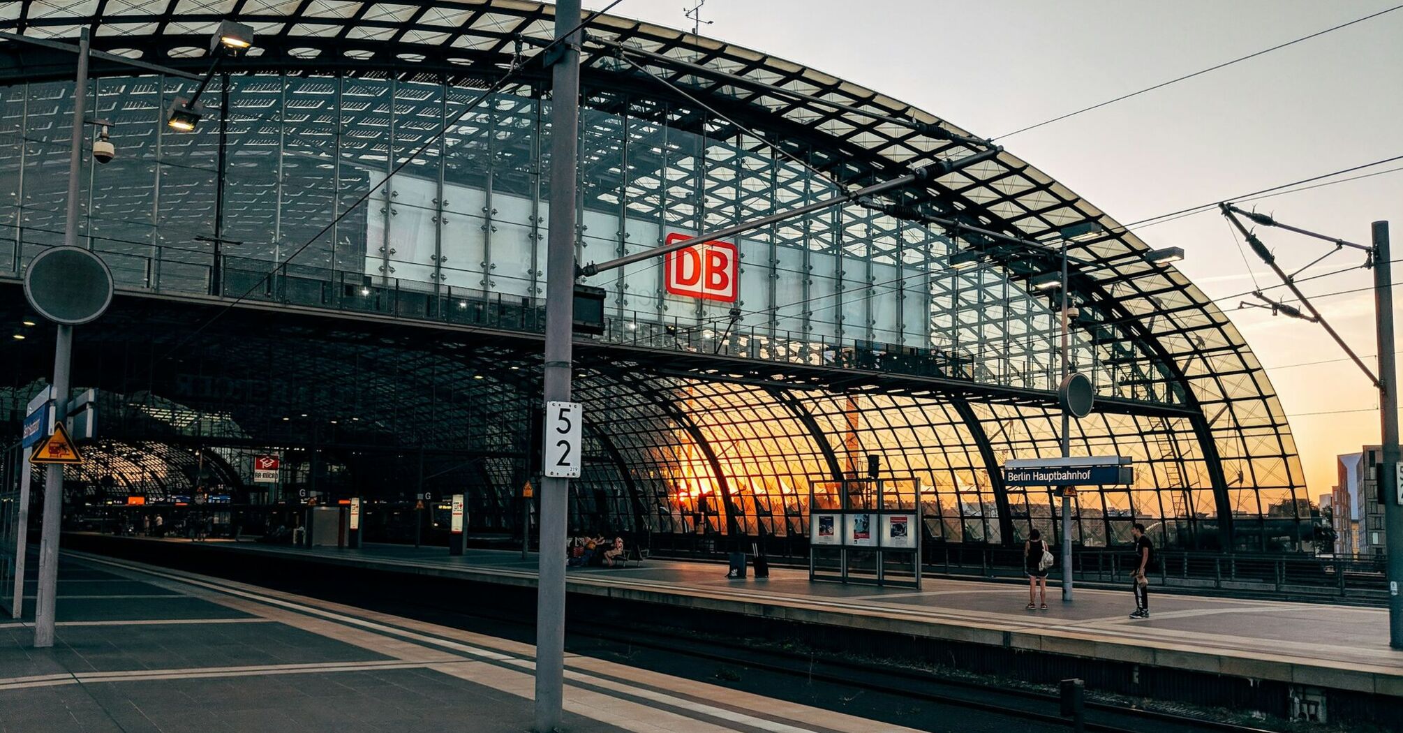 A modern glass-structured railway station with a "DB" sign and a sunset in the background, indicating Berlin Hauptbahnhof