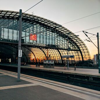 A modern glass-structured railway station with a "DB" sign and a sunset in the background, indicating Berlin Hauptbahnhof