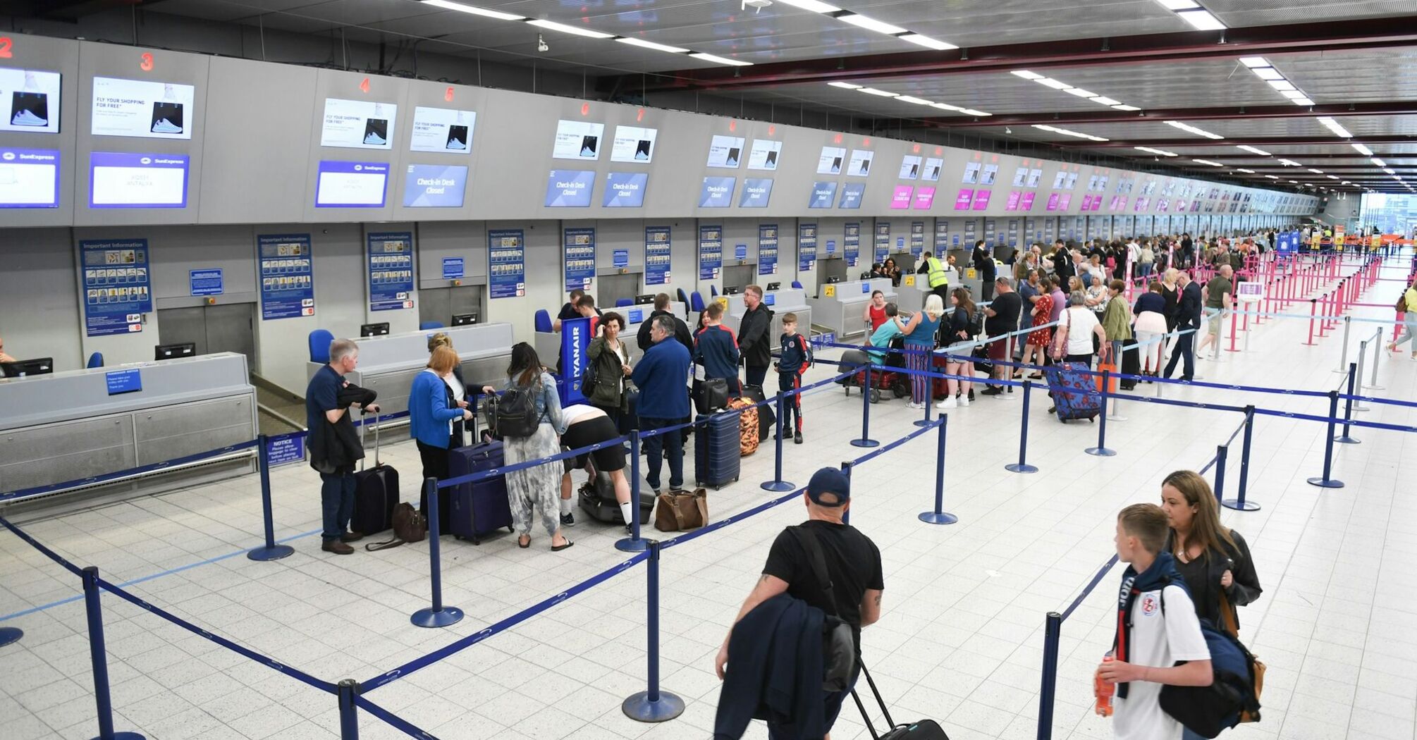 Passengers waiting in line at an airport check-in counter, with luggage and security barriers in view