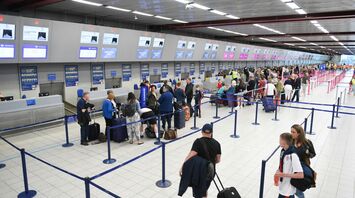 Passengers waiting in line at an airport check-in counter, with luggage and security barriers in view