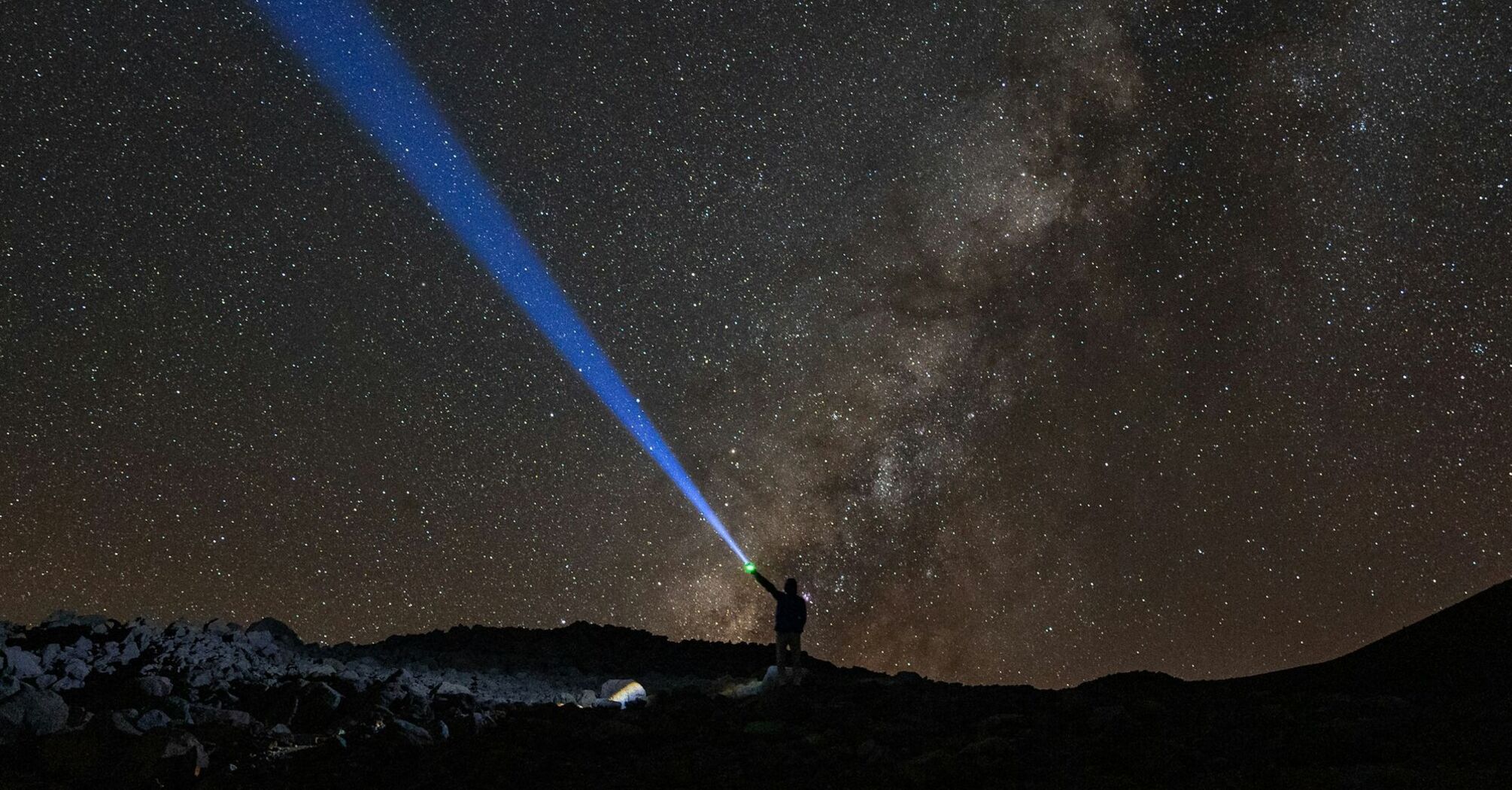 A person shines a blue beam of light into the starry sky over Mauna Kea, Hawaii, with the Milky Way visible in the background