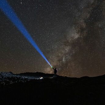 A person shines a blue beam of light into the starry sky over Mauna Kea, Hawaii, with the Milky Way visible in the background