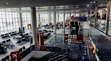 Interior of a modern airport terminal with escalators, gate signs, seating areas, and convenience store signage