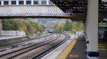 Train approaching a small countryside station with a covered footbridge above the tracks