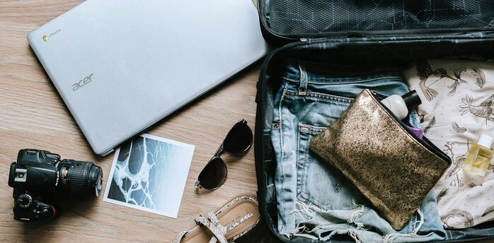 Open suitcase with travel essentials including denim shorts, toiletries, sandals, a camera, and a laptop on a wooden floor