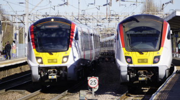 Greater Anglia trains at a station, with a speed limit sign and overhead power lines visible