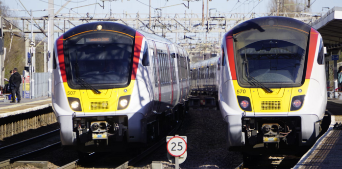 Greater Anglia trains at a station, with a speed limit sign and overhead power lines visible