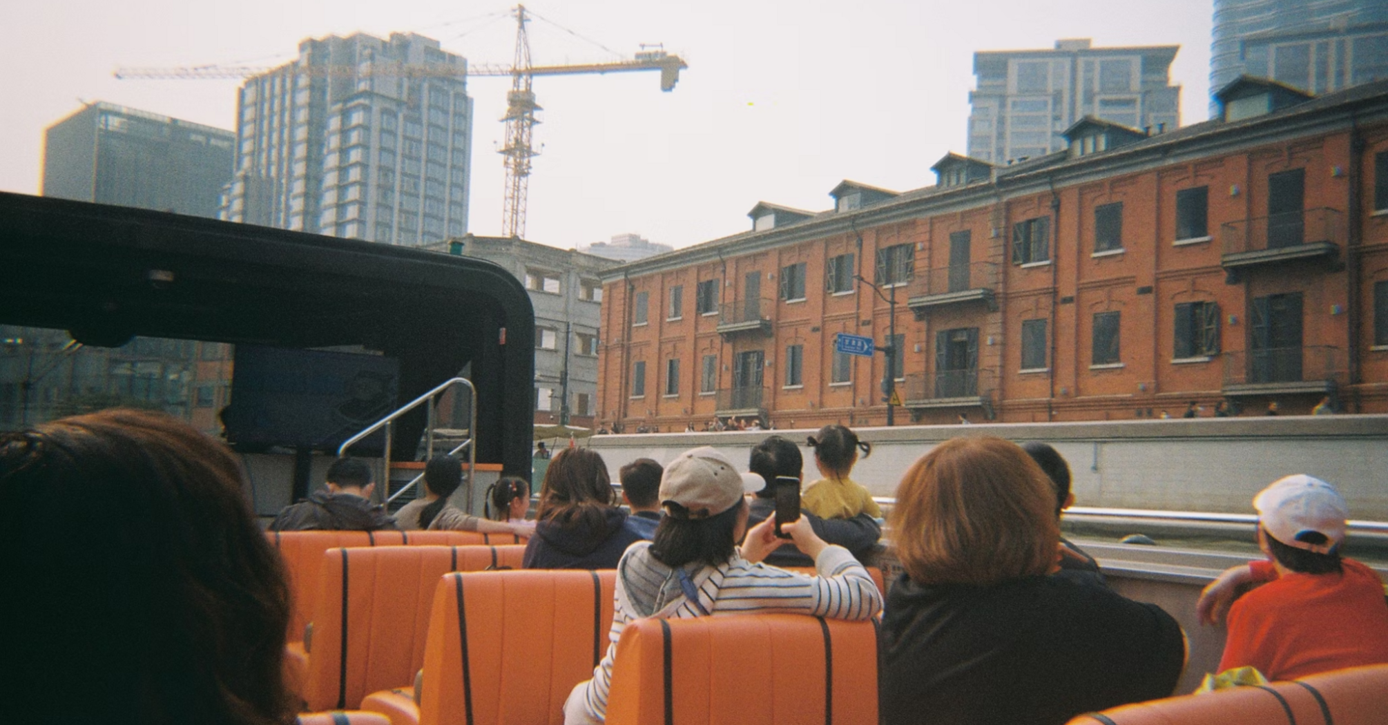 Passengers on an open-top tour bus ride through a city with mid-rise buildings and cranes in the background