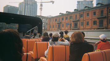 Passengers on an open-top tour bus ride through a city with mid-rise buildings and cranes in the background