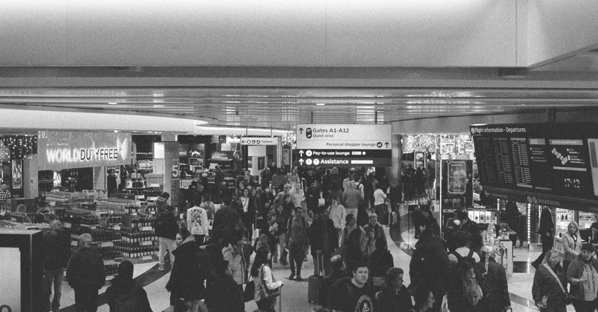 Crowd of passengers in airport terminal with duty-free and flight boards visible