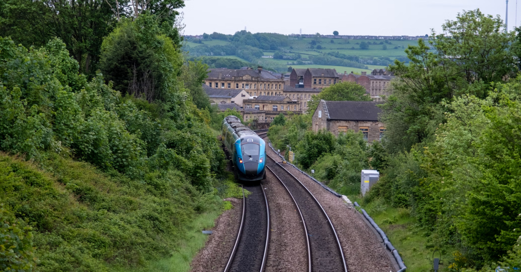 A modern passenger train travels through a rural area in Northern England, surrounded by greenery and village buildings