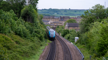 A modern passenger train travels through a rural area in Northern England, surrounded by greenery and village buildings