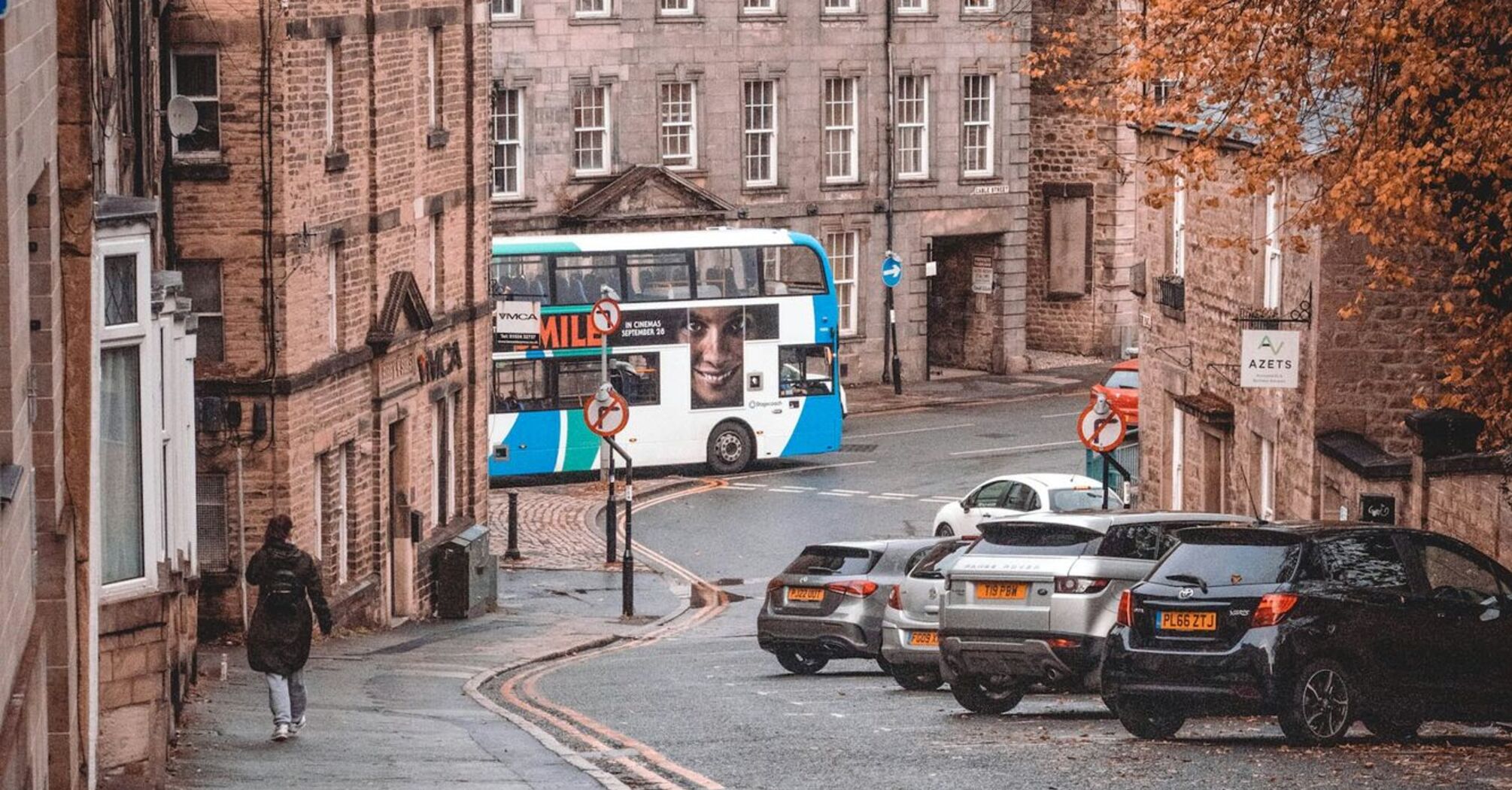 Street view in a historic UK town with a double-decker stagecoach bus, autumn trees, and parked cars
