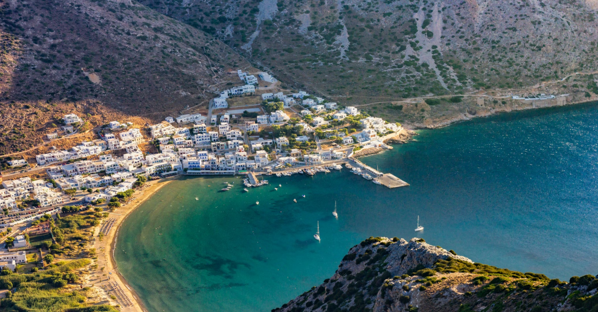 Aerial view of a coastal Greek village surrounded by mountains and a calm turquoise bay