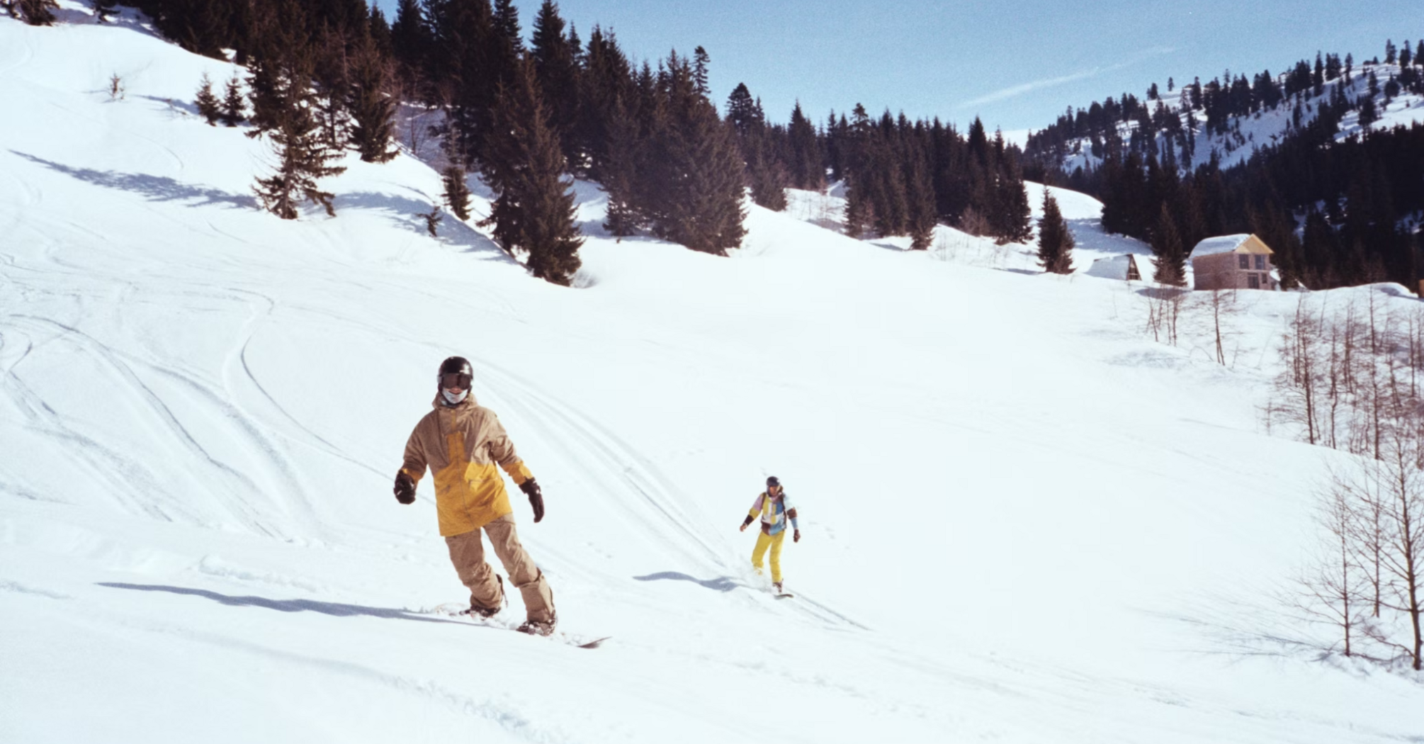 Two people skiing down a snowy mountain slope with pine trees and cabins in the background