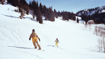 Two people skiing down a snowy mountain slope with pine trees and cabins in the background