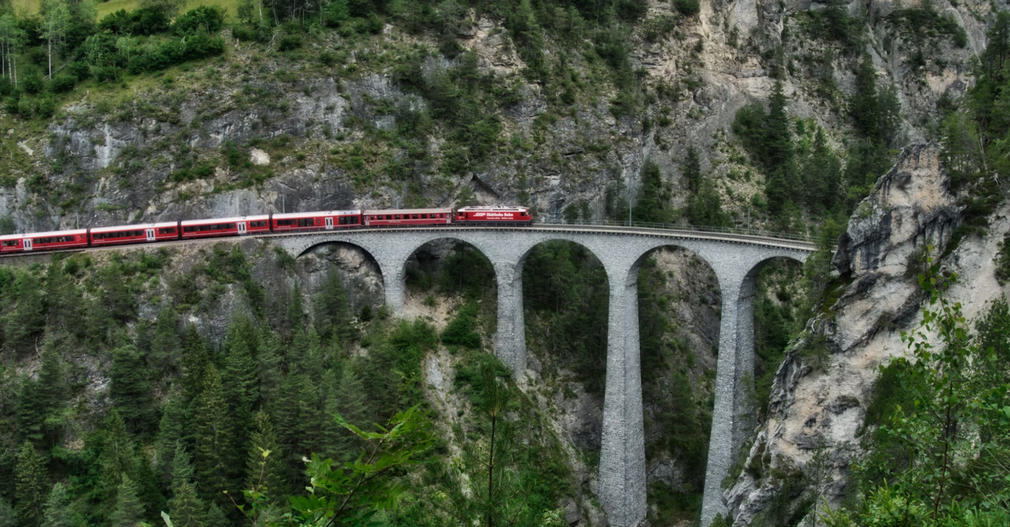 Red passenger train crossing a high stone viaduct in the surrounded by forested cliff