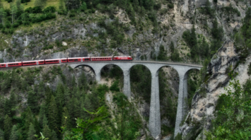 Red passenger train crossing a high stone viaduct in the surrounded by forested cliff