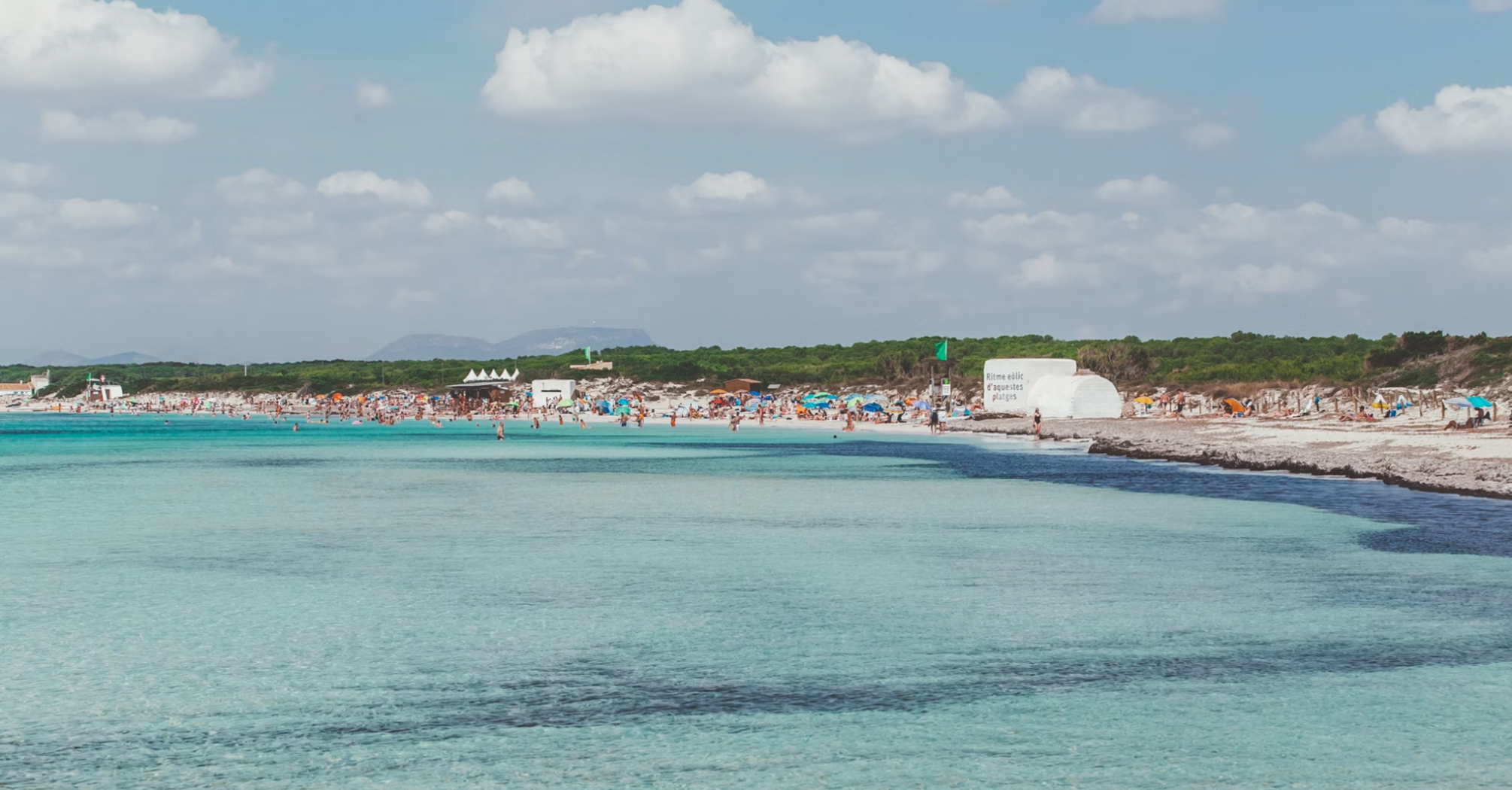 Beachgoers crowd a popular beach on a Balearic Island under a sunny sky