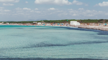 Beachgoers crowd a popular beach on a Balearic Island under a sunny sky