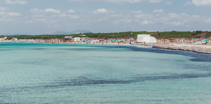 Beachgoers crowd a popular beach on a Balearic Island under a sunny sky