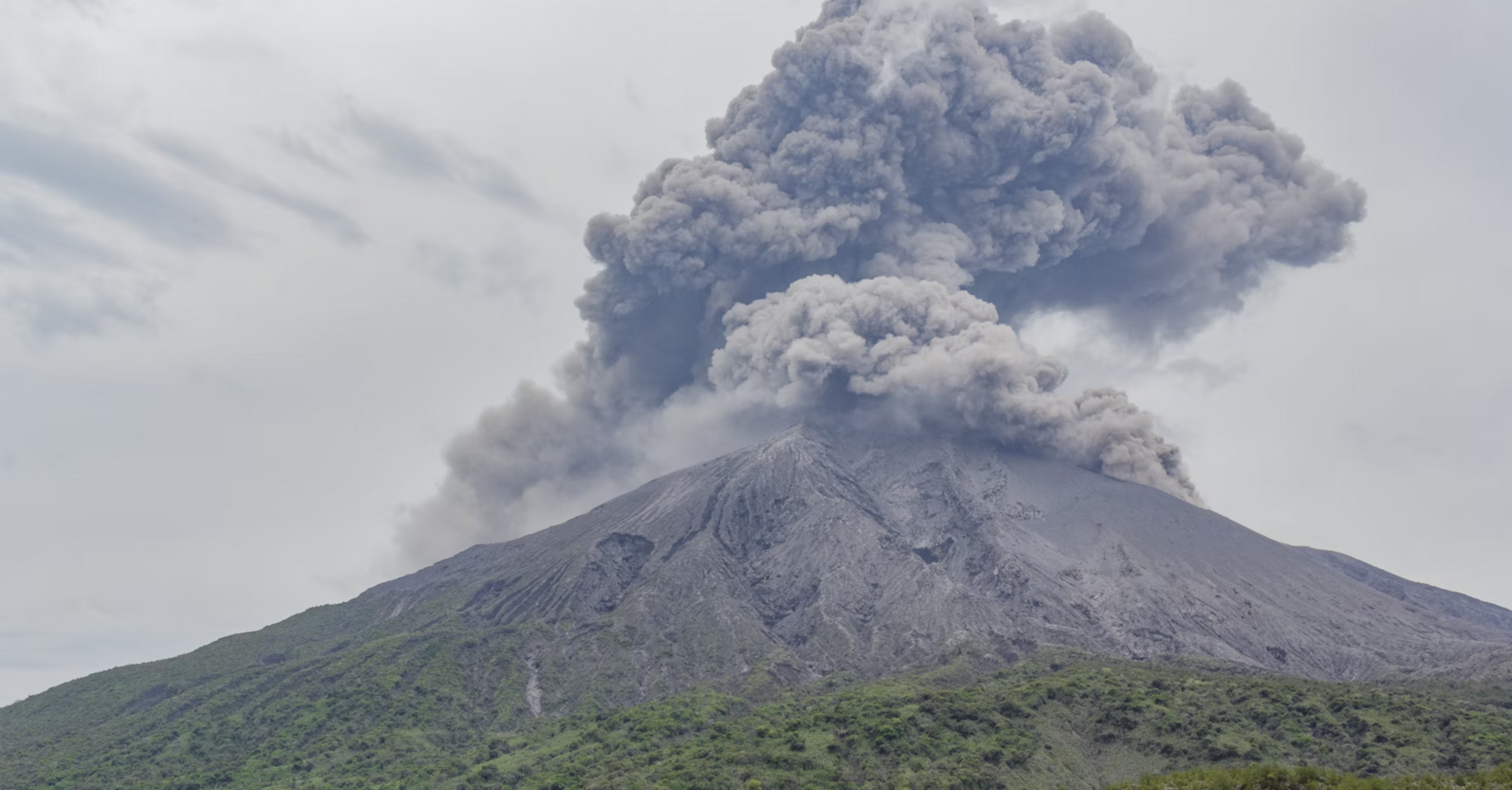 Volcanic eruption sending thick ash clouds into the sky over a green mountainside