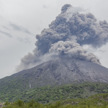 Volcanic eruption sending thick ash clouds into the sky over a green mountainside