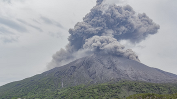 Volcanic eruption sending thick ash clouds into the sky over a green mountainside