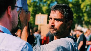 Man holding a protest sign during a daytime demonstration, facing a journalist in a crowd