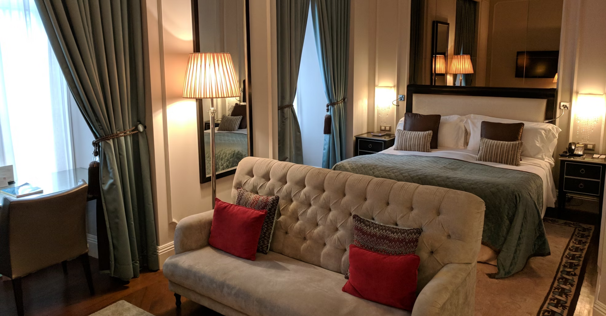 Elegant hotel room with a bed, sofa, and warm lighting, featuring large windows and classic decor