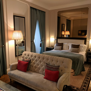 Elegant hotel room with a bed, sofa, and warm lighting, featuring large windows and classic decor
