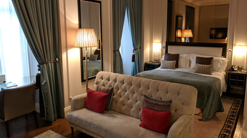 Elegant hotel room with a bed, sofa, and warm lighting, featuring large windows and classic decor