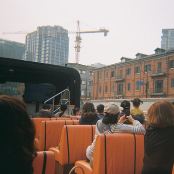 People riding an open-top bus through a city with buildings and cranes in the background