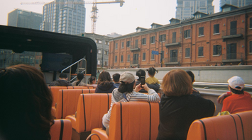 People riding an open-top bus through a city with buildings and cranes in the background