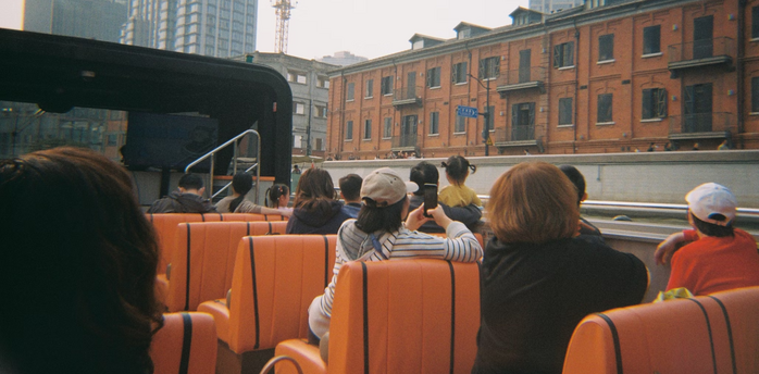 People riding an open-top bus through a city with buildings and cranes in the background