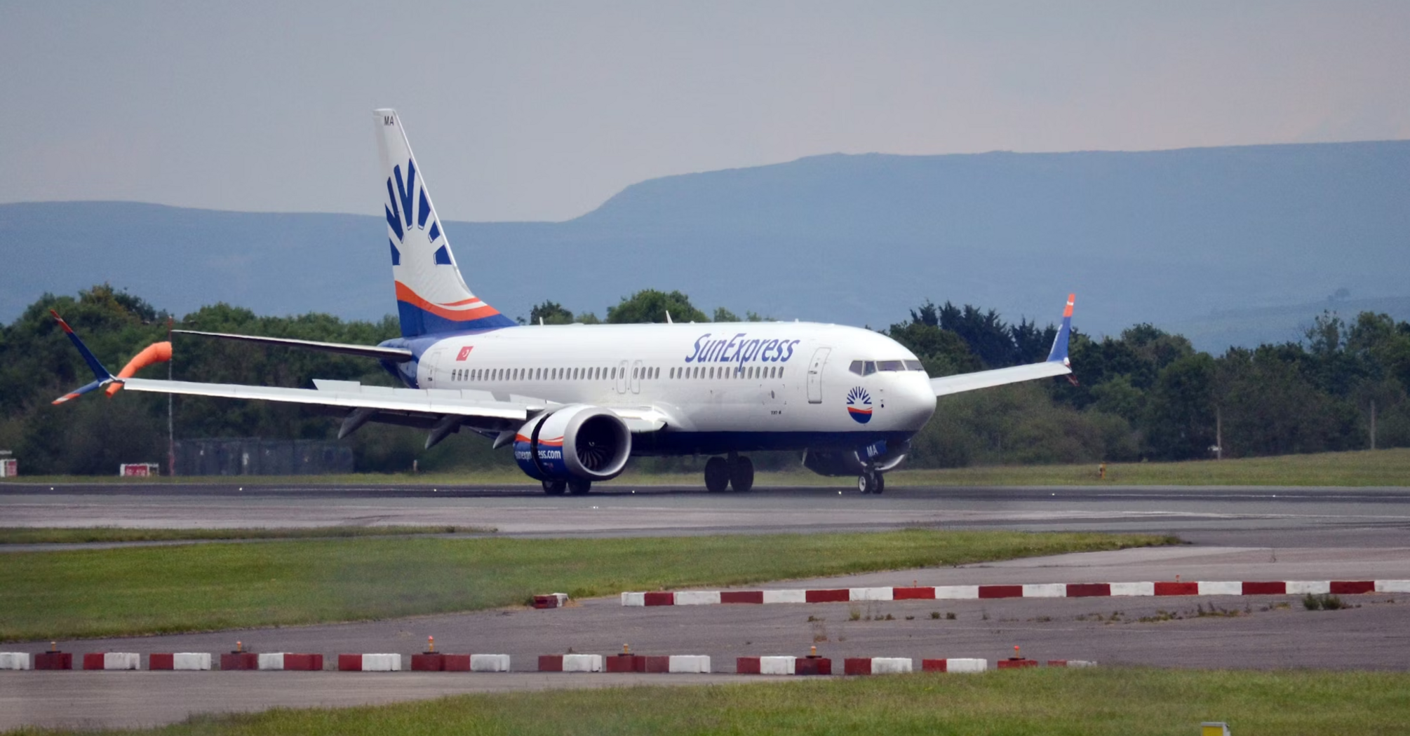 A SunExpress airplane taxiing on the runway at an airport with green hills in the background
