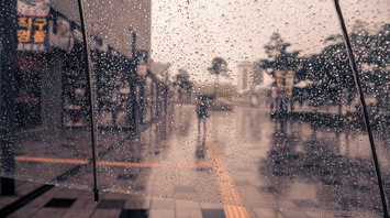 A rainy street scene viewed through a transparent umbrella, with raindrops covering the surface