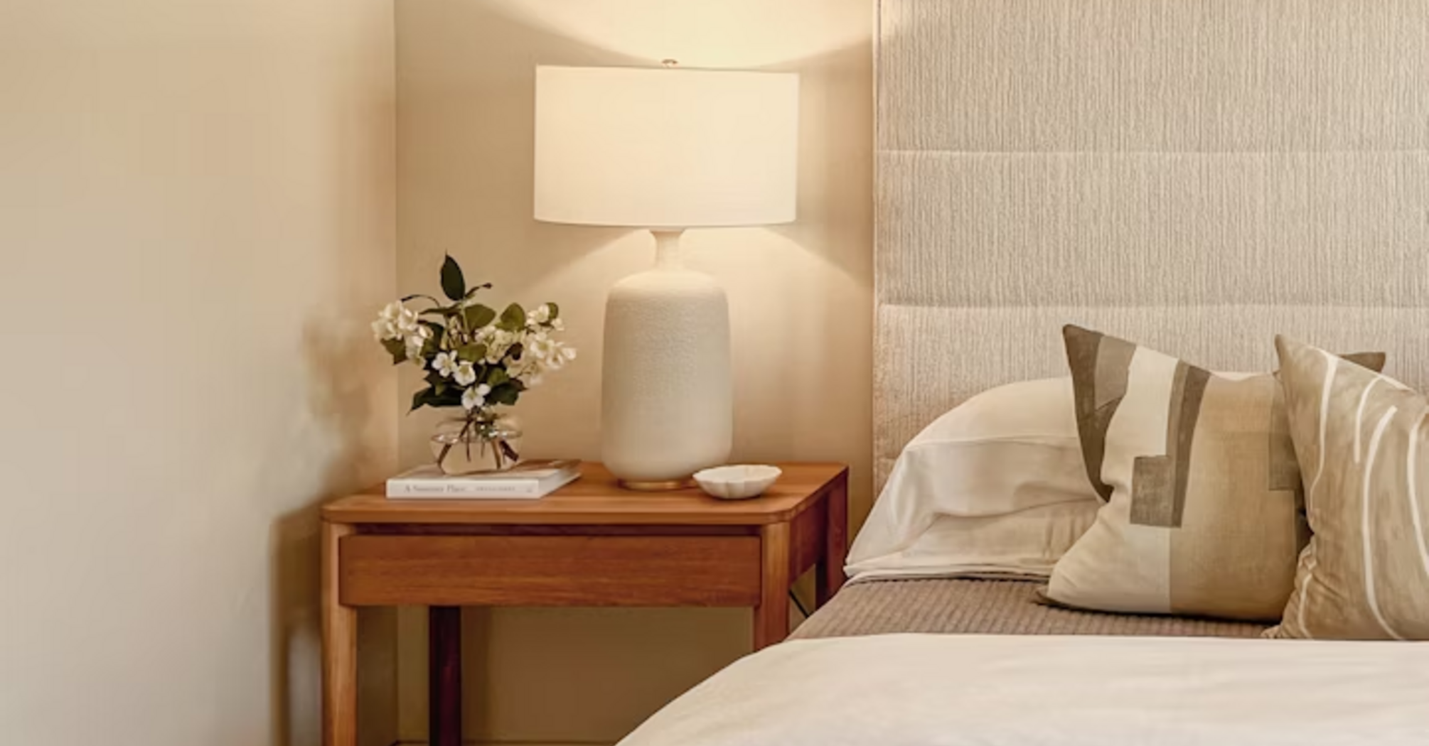 Modern hotel room corner with bedside table, lamp, and neutral bedding